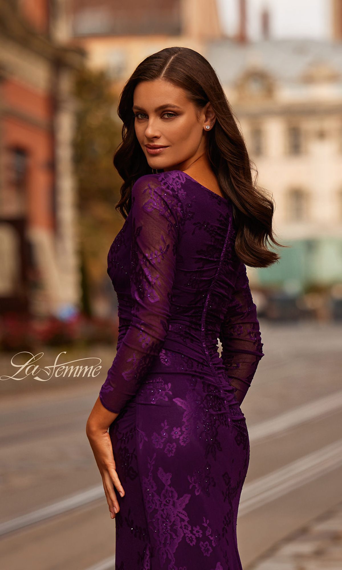 A woman stands outdoors on a city street, looking over her shoulder at the camera in the fitted La Femme 32491 Sheer-Sleeved Lace Formal Dress.